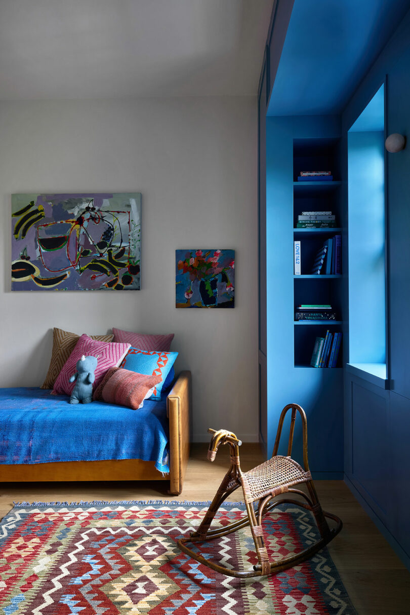 A colorful children's room with a blue bed, patterned rug, wicker rocking horse, blue built-in shelves, and abstract paintings on the wall.