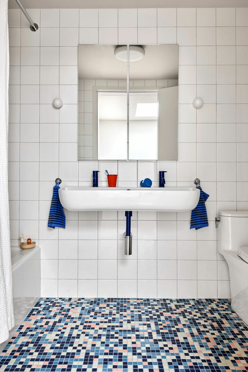 Modern bathroom with white tiled walls, a rectangular sink, two mirrors, blue and white mosaic floor tiles, striped blue towels, and minimal accessories.