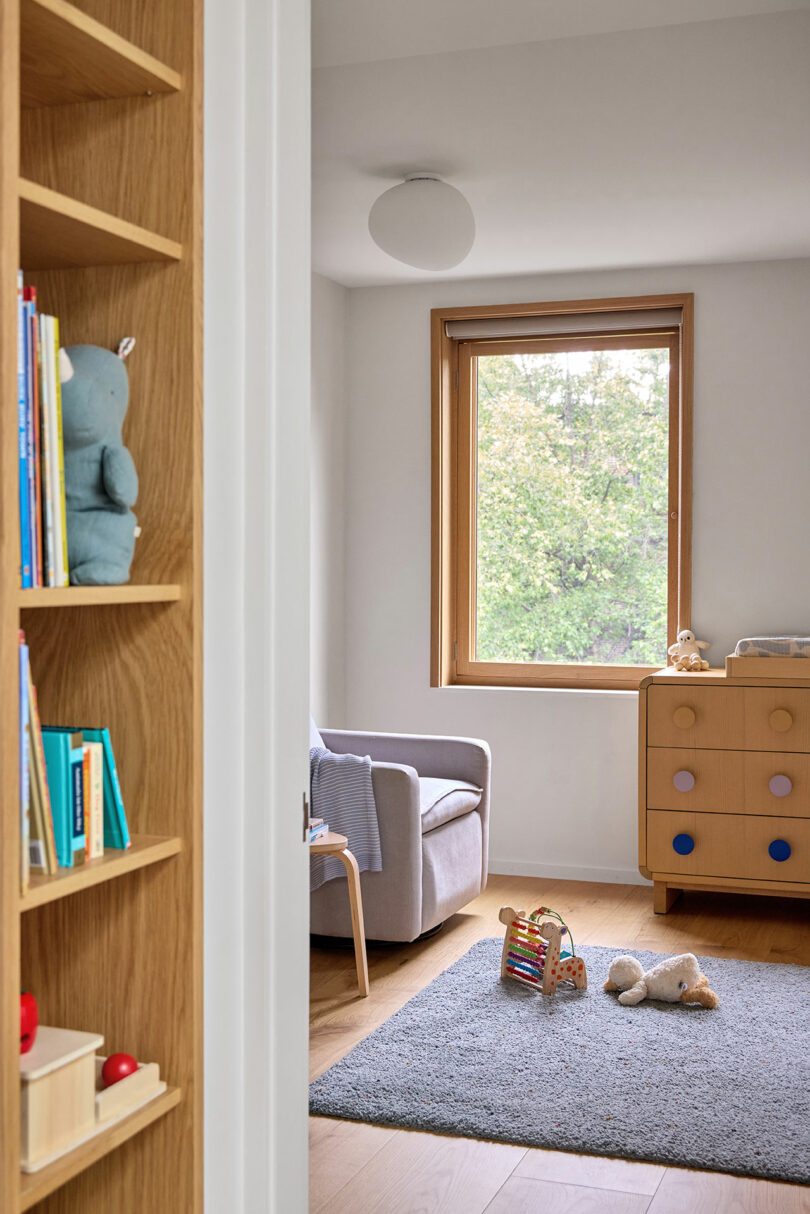 A bright, tidy children's room with a gray armchair, wooden dresser, window, toys on a gray rug, and a bookshelf partially visible in the foreground.