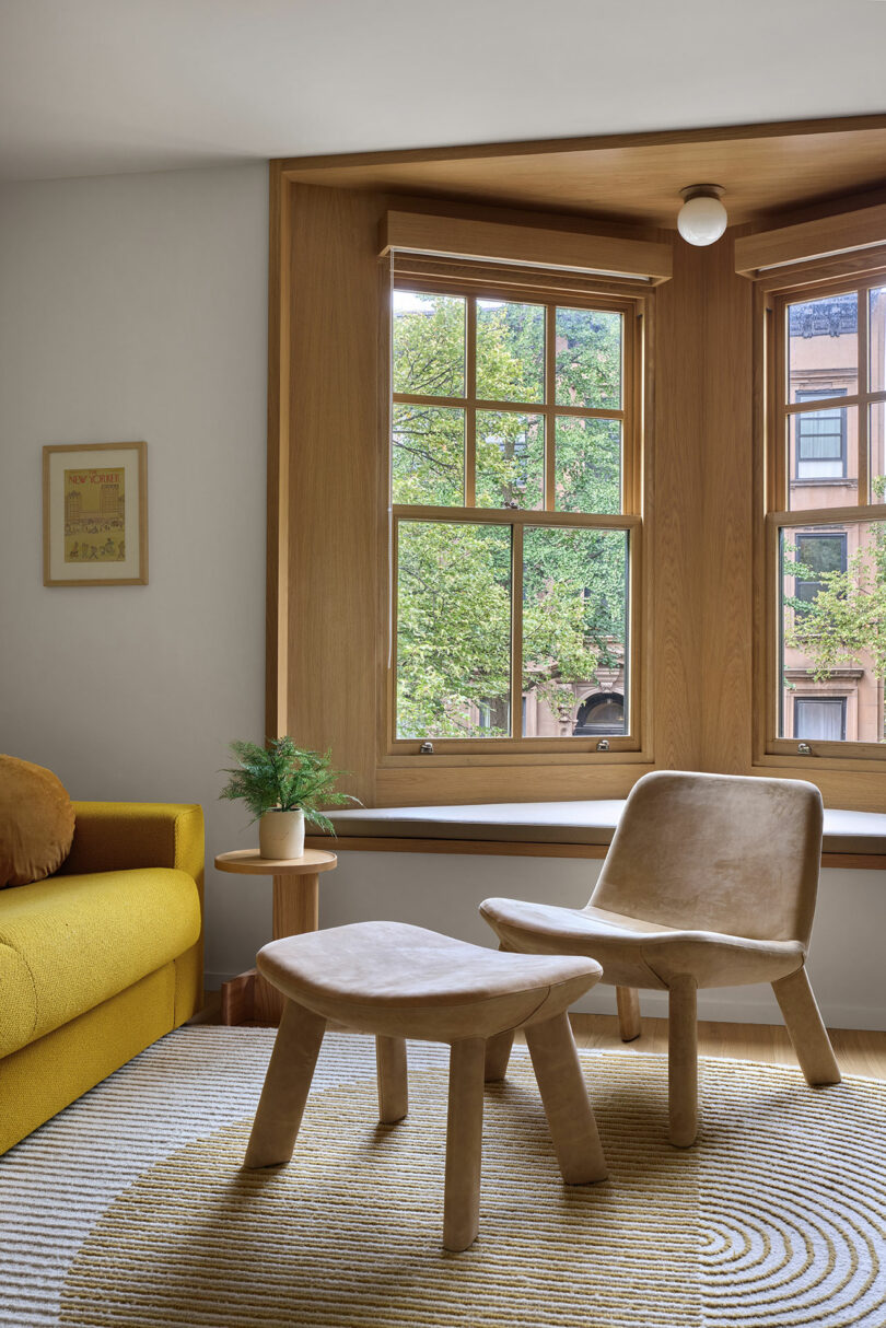 A cozy reading nook with wooden chairs, a small side table, and a window seat overlooks leafy trees through large bay windows. A yellow sofa is visible on the left.