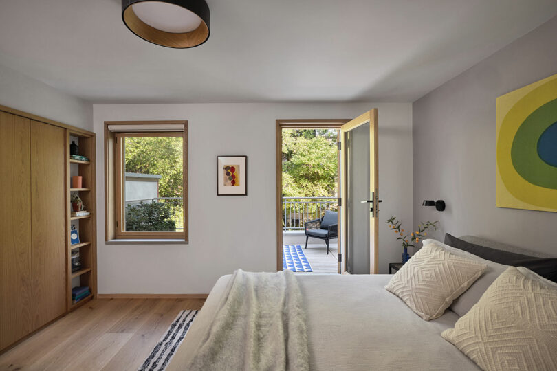 A modern bedroom with a neatly made bed, wooden floors, built-in shelves, and an open door leading to a deck with outdoor seating.
