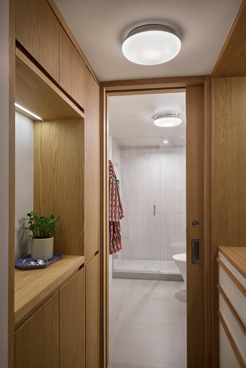 A hallway with built-in wood cabinetry leads to a bathroom with a glass shower, tiled walls, and a checked robe hanging on the wall.
