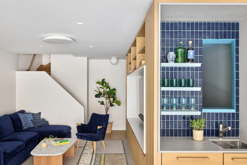 Modern living room with a navy blue sectional sofa, wooden chair, marble coffee table, and a kitchenette area with blue tile, open shelves, and a small sink.