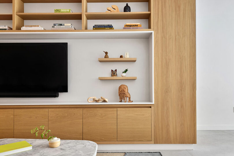 Modern living room with built-in wooden shelves and cabinets, a wall-mounted TV, decorative items on floating shelves, and a marble coffee table with a small plant.