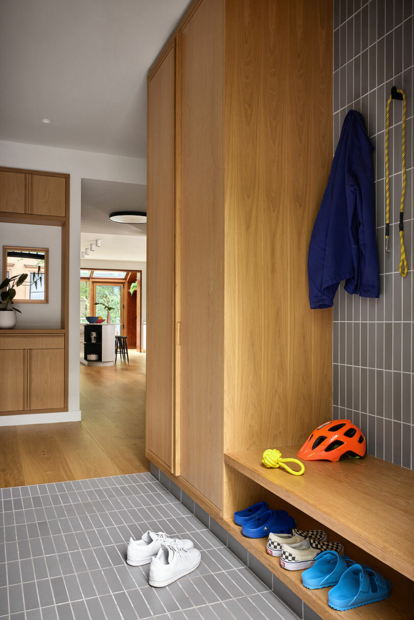 A modern mudroom with wooden cabinets, gray tile flooring, bench, shelves with shoes, wall hooks holding a blue jacket, orange helmet, and dog leash.