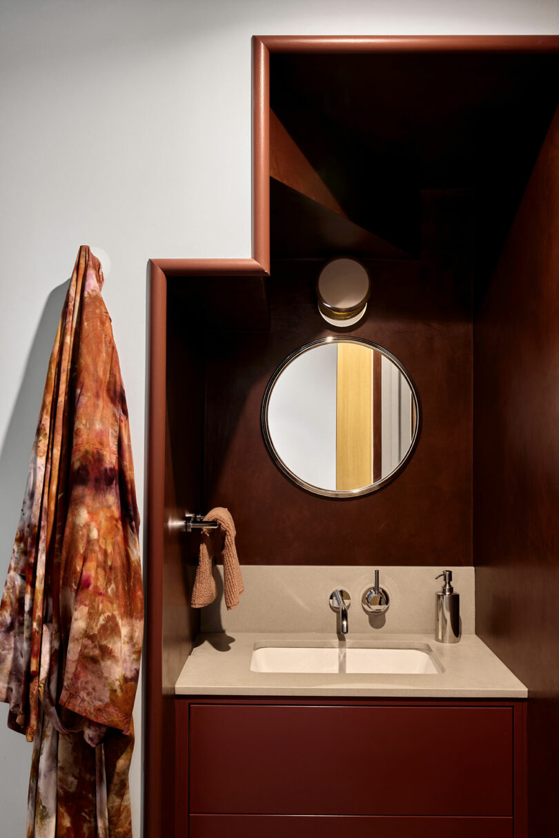 A modern bathroom sink with a circular mirror, wall-mounted faucet, small towel, soap dispenser, and a colorful robe hanging on a hook.