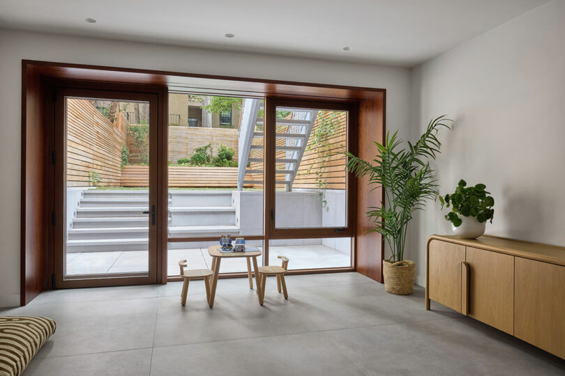A modern room with large glass doors opening to a terraced backyard, featuring a small wooden table with stools, plants, and a light wood sideboard.