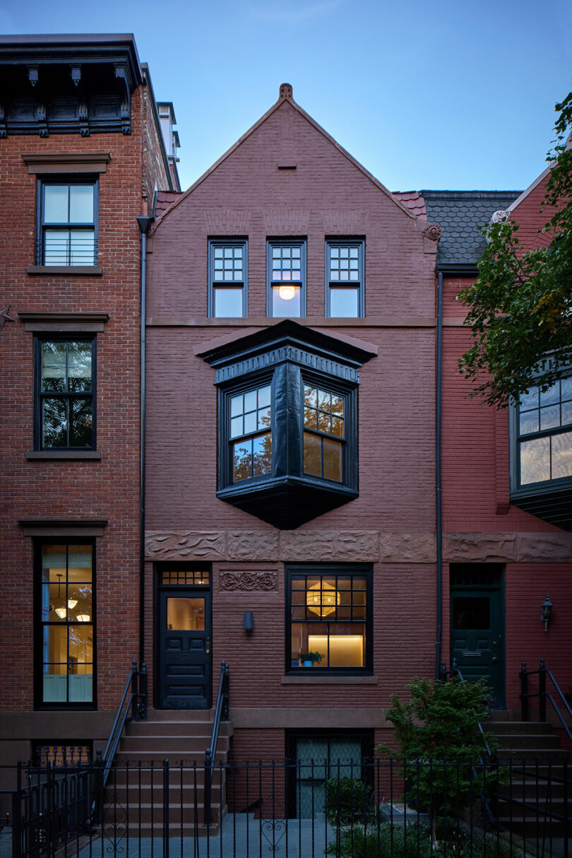 A brick townhouse with a bay window, black door, front steps, and iron fence, flanked by similar neighboring buildings at dusk.