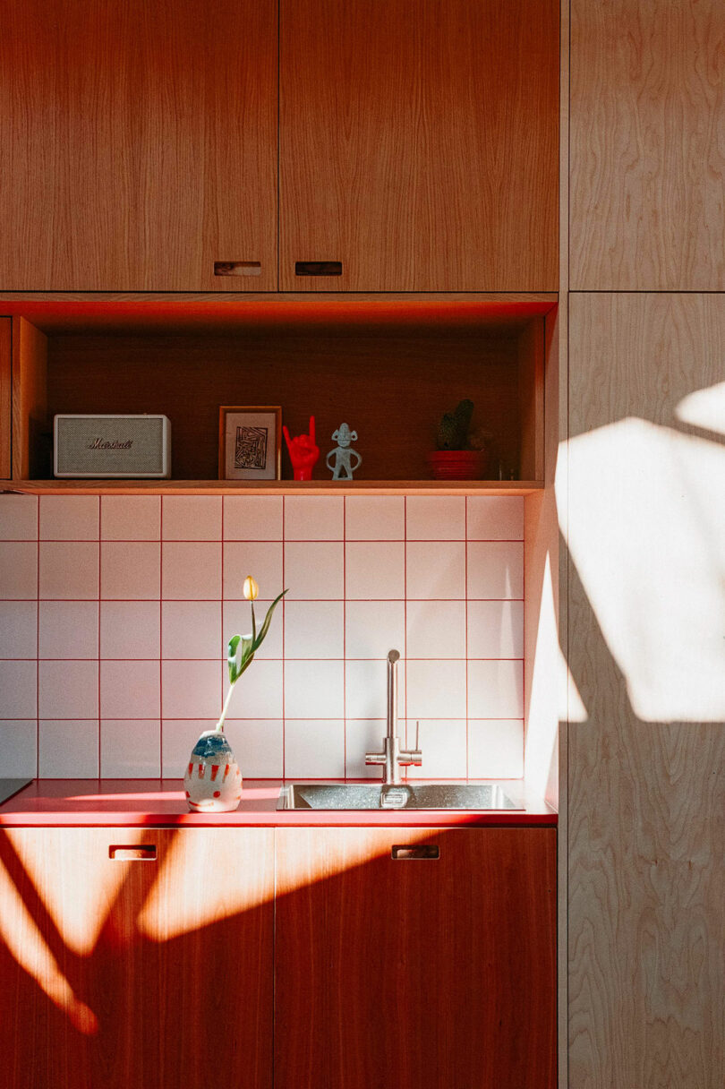 Modern kitchen sink with a single flower in a vase on the counter, wooden cabinets, open shelving, and sunlight casting shadows on the tiled backsplash and wall.