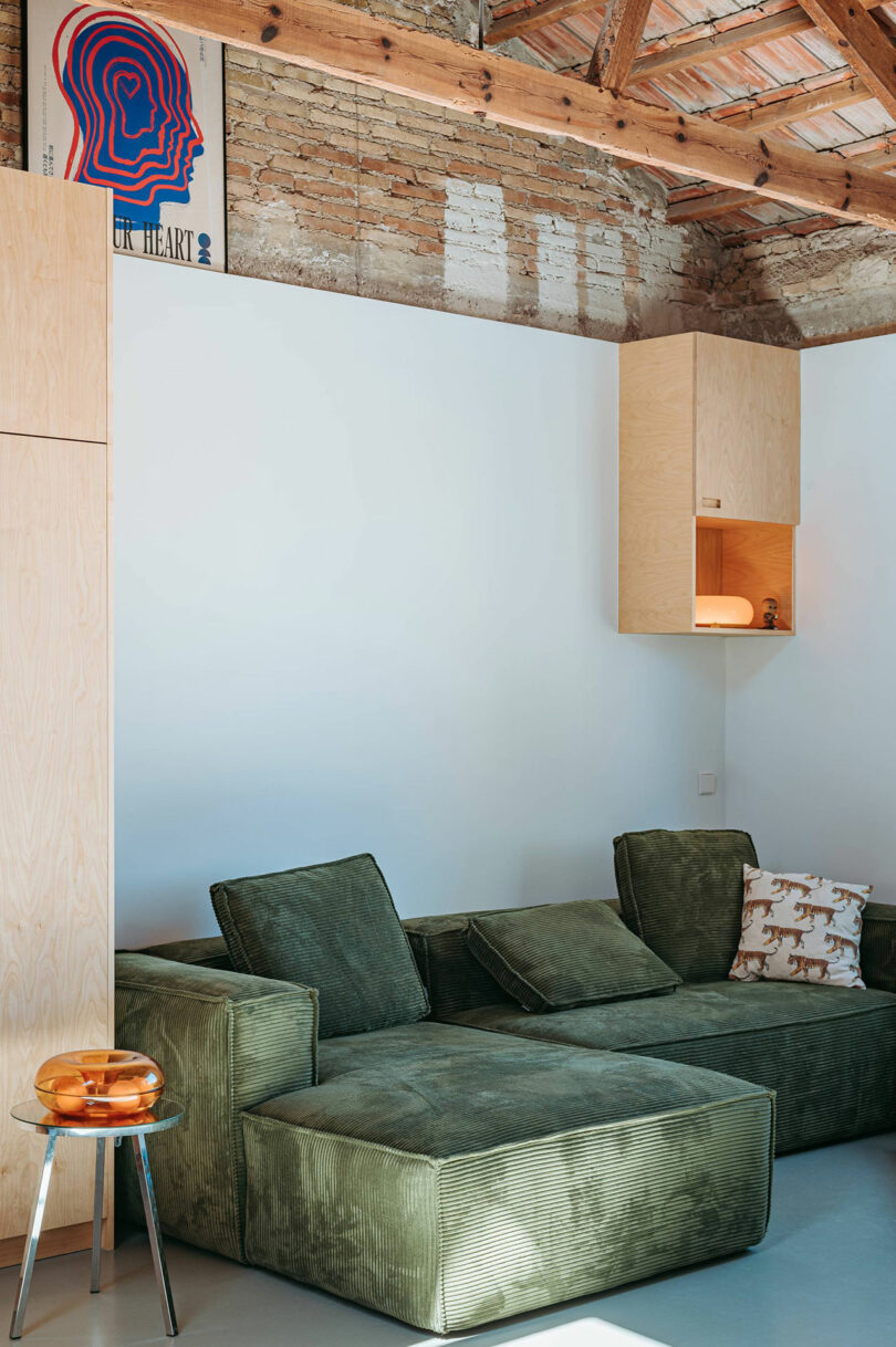 A green sectional sofa with patterned cushions sits in a modern living room with exposed brick, wooden ceiling beams, and minimal decor, including a small side table with amber glass objects.