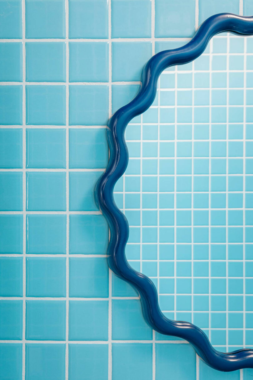 A wavy-edged blue mirror partially reflects light blue square tiles on a bathroom wall.