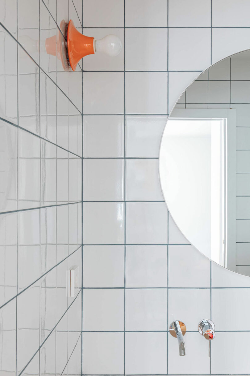 White tiled bathroom wall with a round mirror reflecting a doorway, an exposed light bulb with an orange fixture, and two metal taps below the mirror.