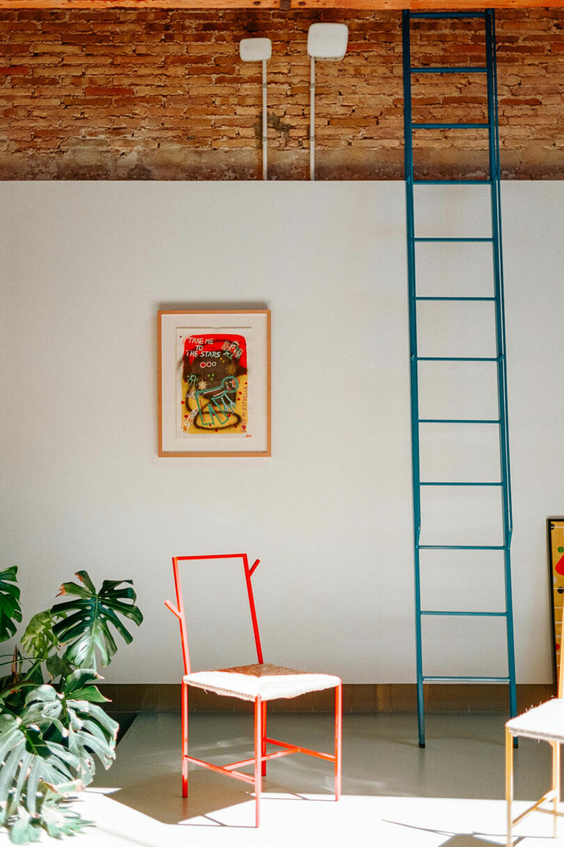 A minimalist room features a red chair, a modern art piece on a white wall, a blue metal ladder leading up, and green plants in the corner. Exposed brick is visible at the top.