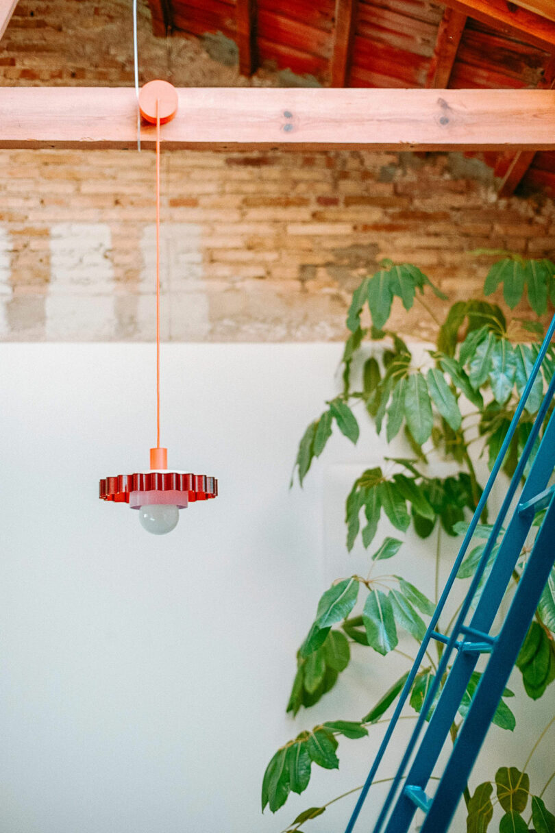 An orange pendant light hangs from a wooden beam ceiling near a green potted plant and a blue metal staircase against a white wall.