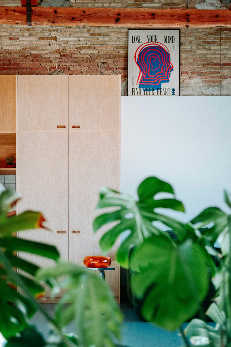 Modern interior with light wood cabinets, exposed brick wall, and a colorful poster reading “Lose Your Mind, Find Your Heart”; green plant leaves in the foreground.