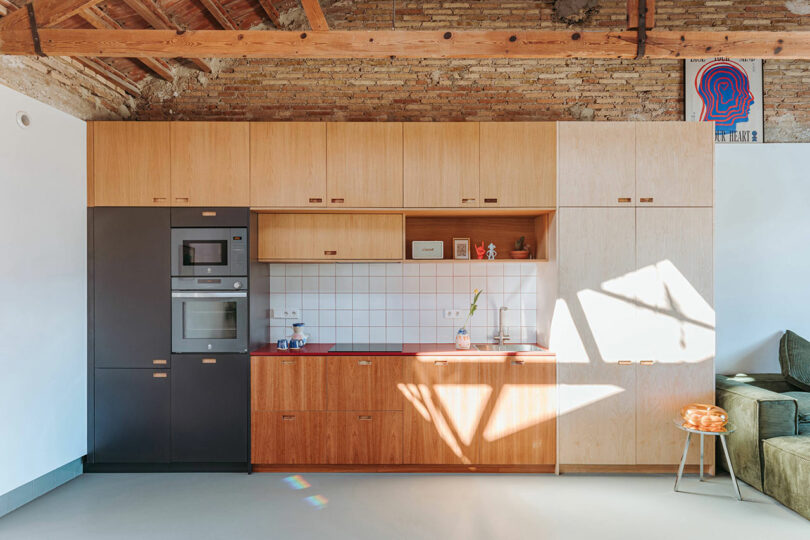 Modern kitchen with wood cabinets, built-in appliances, and white tile backsplash; sunlight casts geometric shadows on the floor and cabinets; exposed brick and wood beam ceiling.