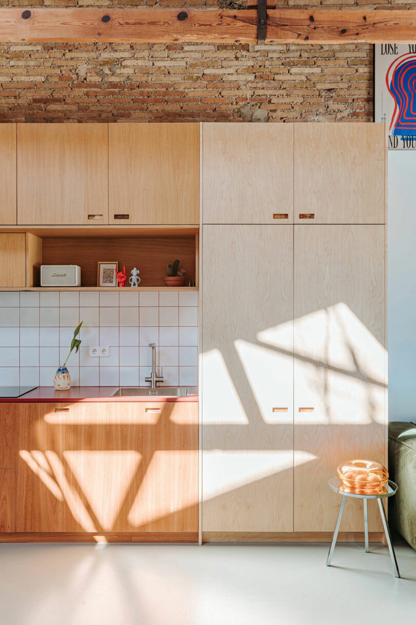 Modern kitchen with light wood cabinets, white tile backsplash, and exposed brick beam. Sunlight casts geometric shadows across the cabinets and a stool with oranges sits beside a green sofa.