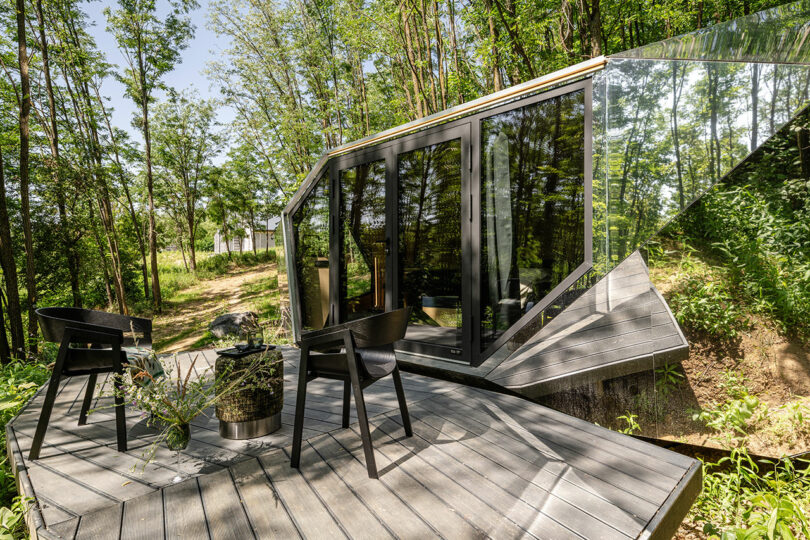 Modern cabin with large glass windows and angular design, set in a wooded area, featuring a small deck with two chairs and a table.