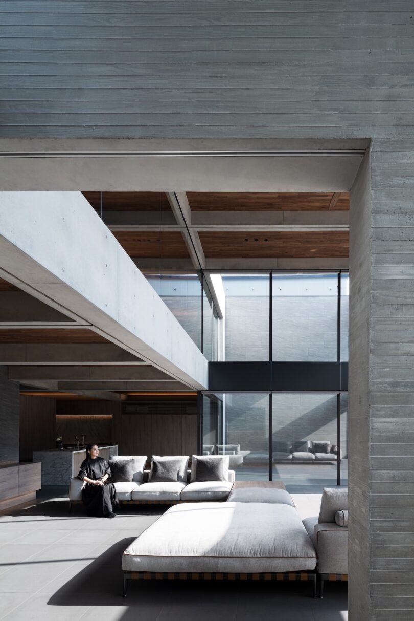 A spacious modern living room with concrete walls, large windows, a sectional sofa, and a person sitting at one end.