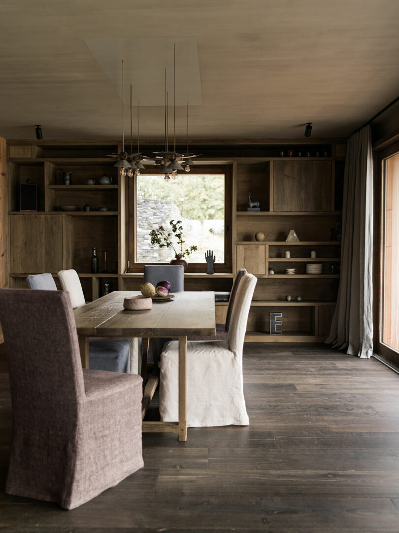 A modern dining room with a wooden table, six fabric-covered chairs, built-in wooden shelves, decorative items, and large windows letting in natural light.