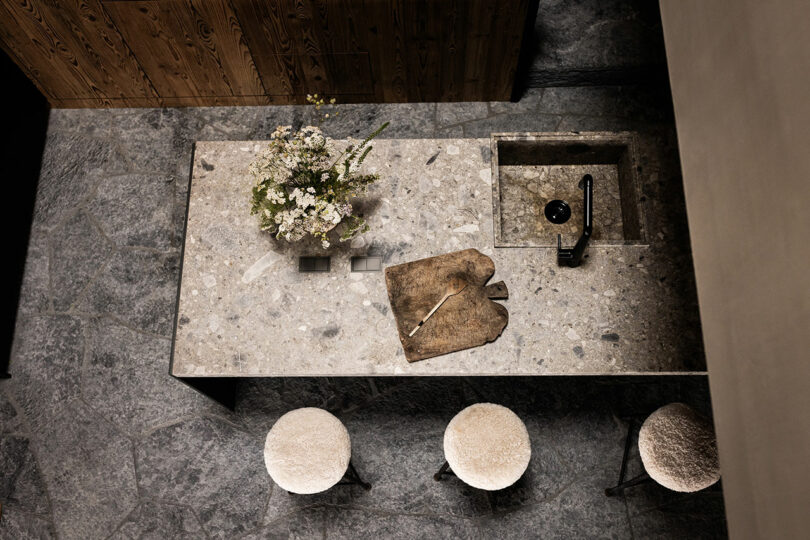 Overhead view of a stone kitchen island with a sink, a cutting board, knife, flower vase, and three round stools on a textured stone floor.