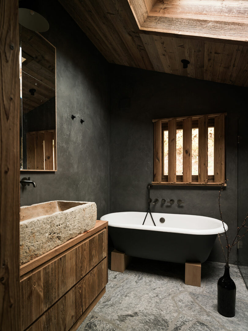 A modern rustic bathroom with a stone sink, freestanding bathtub, wood accents, slate floor, and a skylight.