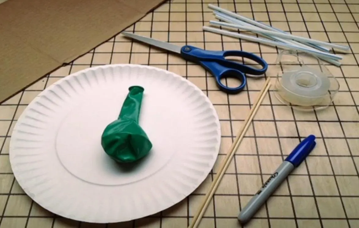 Crafting materials including scissors, a green balloon, a paper plate, and straws on a grid-patterned surface.
