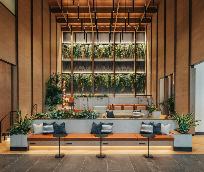 Modern lobby with high ceilings, a vertical garden wall, long bench seating with cushions, and plants—perfect for unwinding after a Friday Five. Warm lighting enhances the wood decor in the central lounge area with sofas and armchairs.