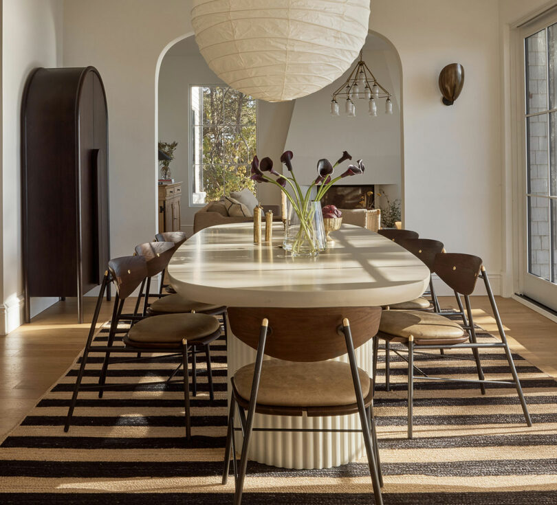 A modern dining room with a long oval table, eight chairs, a striped rug, and a large paper lantern light creates the perfect Friday Five vibe. A vase with dark flowers sits in the center as sunlight streams in through glass doors.