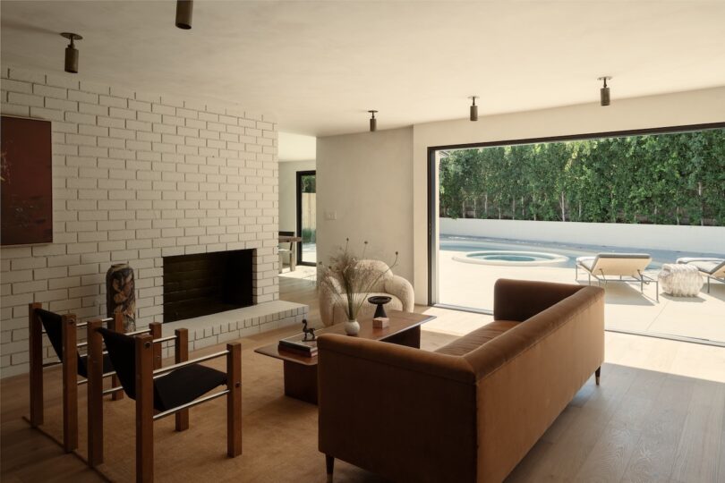 A modern living room with minimalist furniture in soothing skim milk tones, a brick fireplace, and large sliding doors opening to a patio with lounge chairs, a pool, and lush greenery.