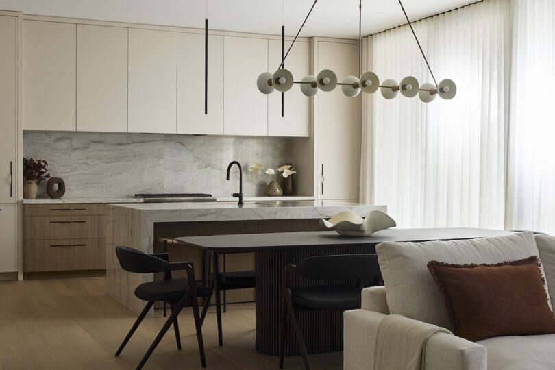 Modern kitchen and dining area with minimalist cabinetry, marble backsplash, black fixtures, pendant lighting, and neutral-toned furniture. Soft daylight—reminiscent of skim milk—filters through sheer curtains.