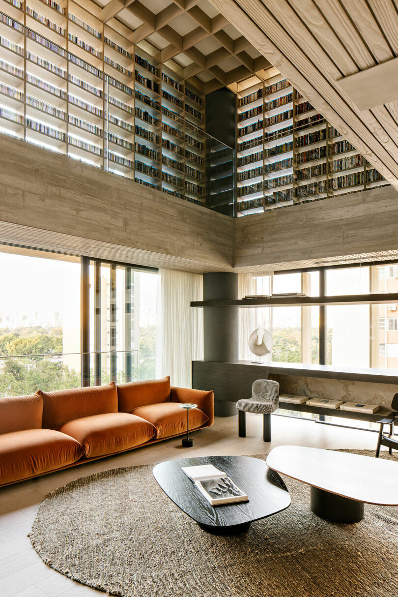 Modern living room with an orange sofa, round rug, black coffee tables, large windows, and a double-height ceiling featuring a lofted wall of bookshelves.