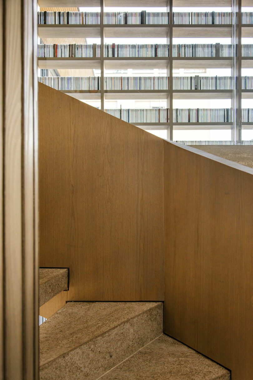Interior view of a staircase with wooden paneling and stone steps; in the background, a large window reveals rows of bookshelves.