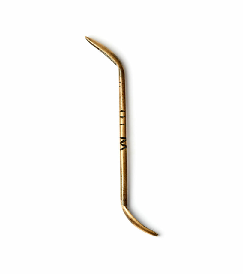 A gold double-ended metal tool with curved tips at both ends, the Brent Buck tool is displayed against a plain white background.