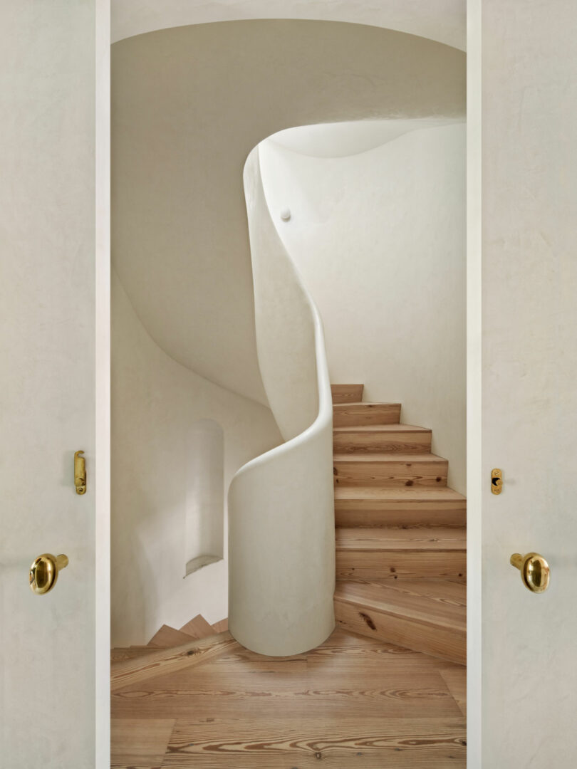 A modern spiral staircase by Brent Buck features light wooden steps and smooth white plastered walls, viewed through an open doorway with brass handles.