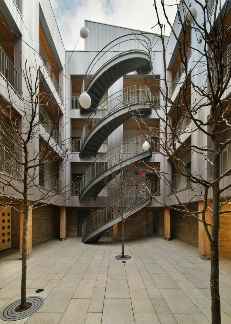 A modern courtyard by Brent Buck with bare trees and a central spiral staircase leading to multiple floors of an apartment building. Spherical white lights are suspended above.