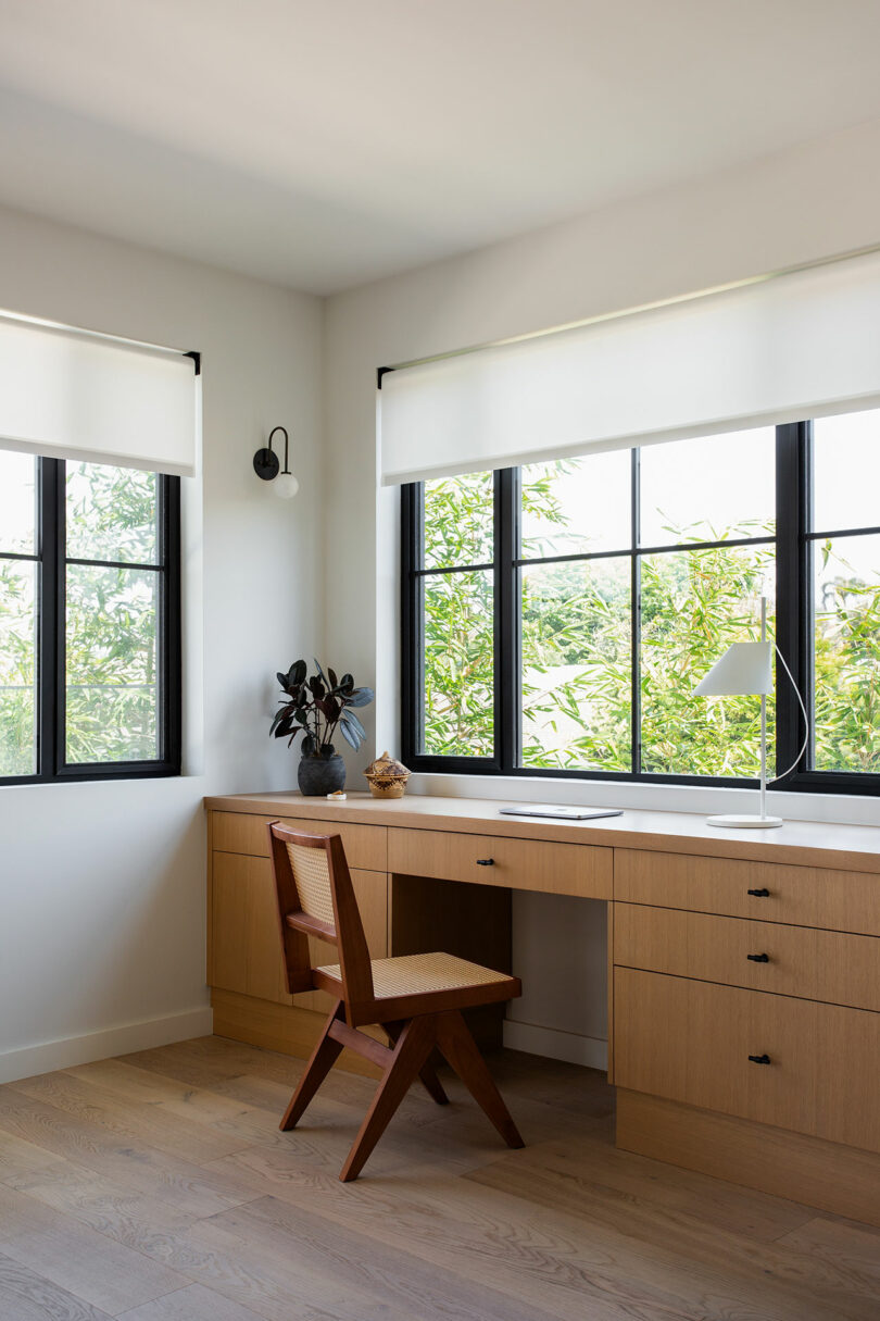 Minimalist home office with a wooden desk and chair, drawers, a white lamp, a potted plant, and large windows with white blinds letting in natural light.