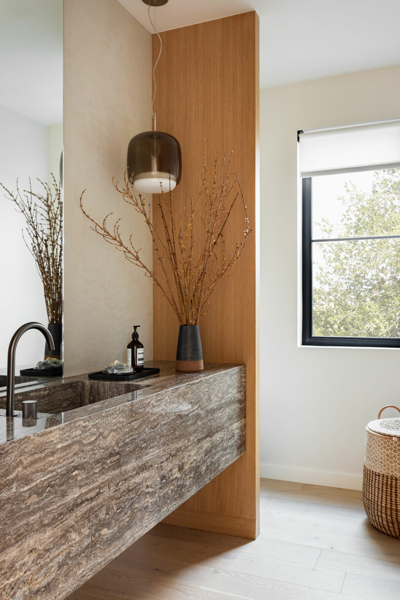 Modern bathroom with a stone sink, wall-mounted faucet, pendant light, tall vase with branches, large mirror, and a window next to a woven basket.