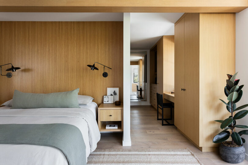 Modern bedroom with wood panel walls, a neatly made bed, wall-mounted lights, built-in desk area, and a potted plant on light wood flooring.