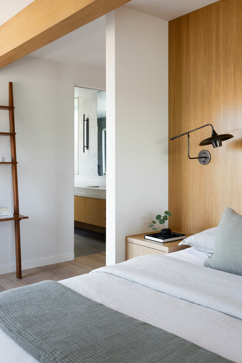 A modern bedroom with wood accents, neatly made bed, wall-mounted lamp, and a view into a minimalist bathroom with light cabinetry.