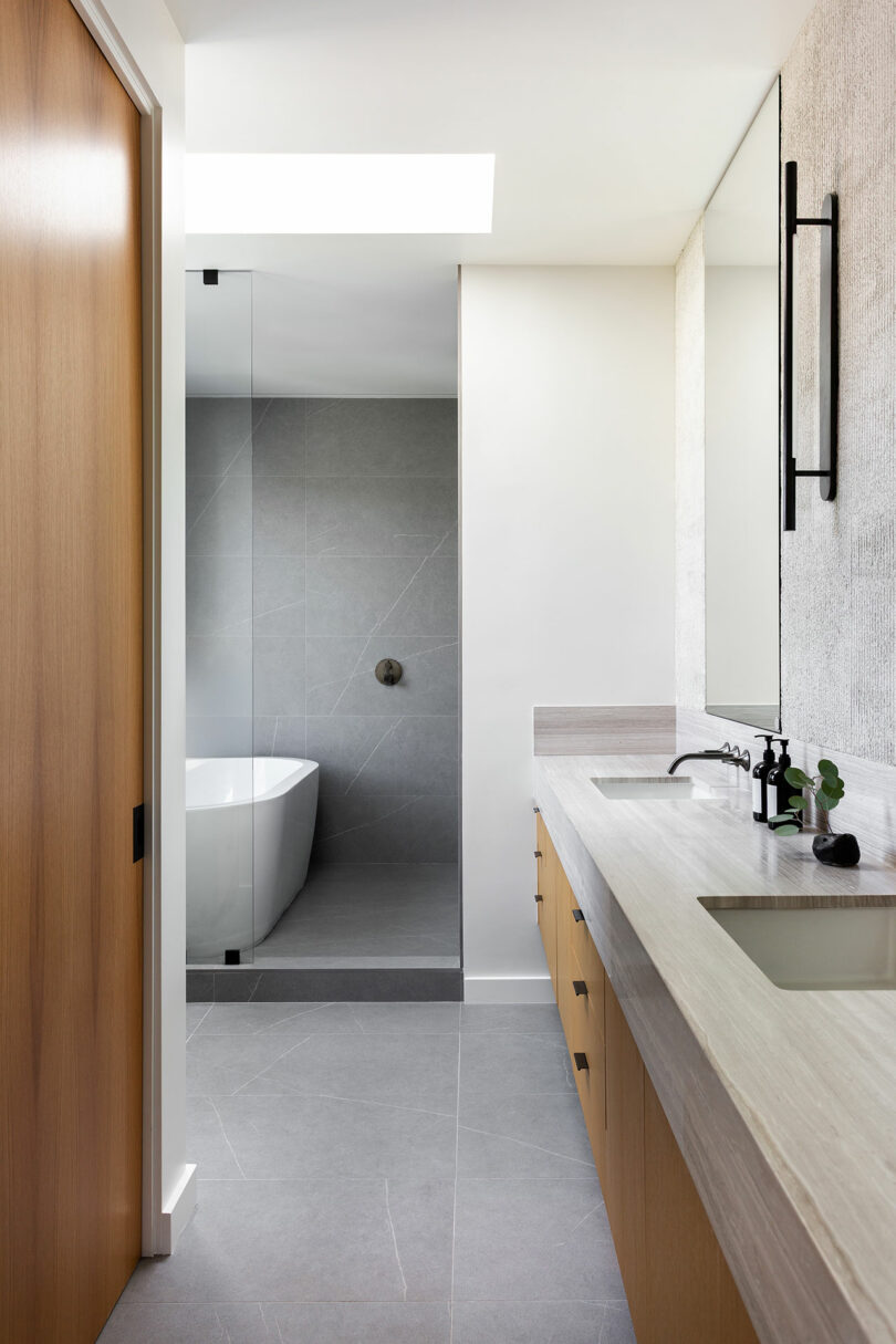 Modern bathroom with light wood cabinetry, large mirror, rectangular sink, and walk-in shower with glass door and freestanding bathtub. Neutral gray and white tones throughout.