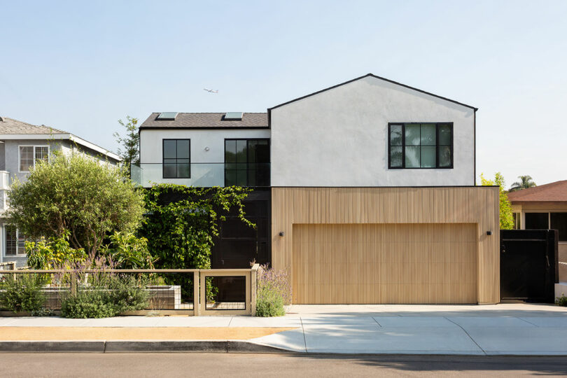 A modern two-story house with a mix of white stucco and light wood exterior, large windows, a double garage, and a small landscaped front yard.
