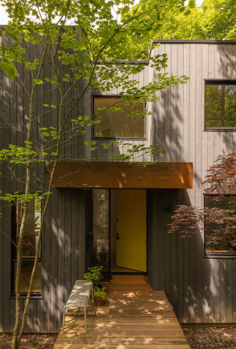 A modern house exterior with vertical wooden siding, large black-framed windows, and a rust-colored awning above a glass door, surrounded by trees and greenery.