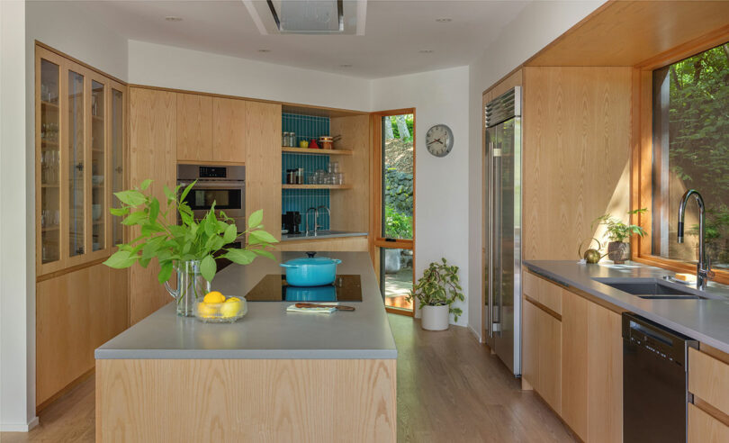 Modern kitchen with light wood cabinets, gray countertops, a center island with a plant and lemons, open shelving, and large windows letting in natural light.