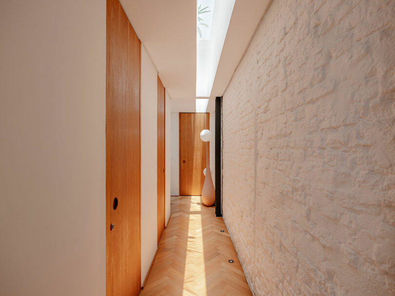 Bright hallway with wood doors, a textured white stone wall, light wood herringbone floor, and a tall white floor lamp, illuminated by natural light from a skylight above.