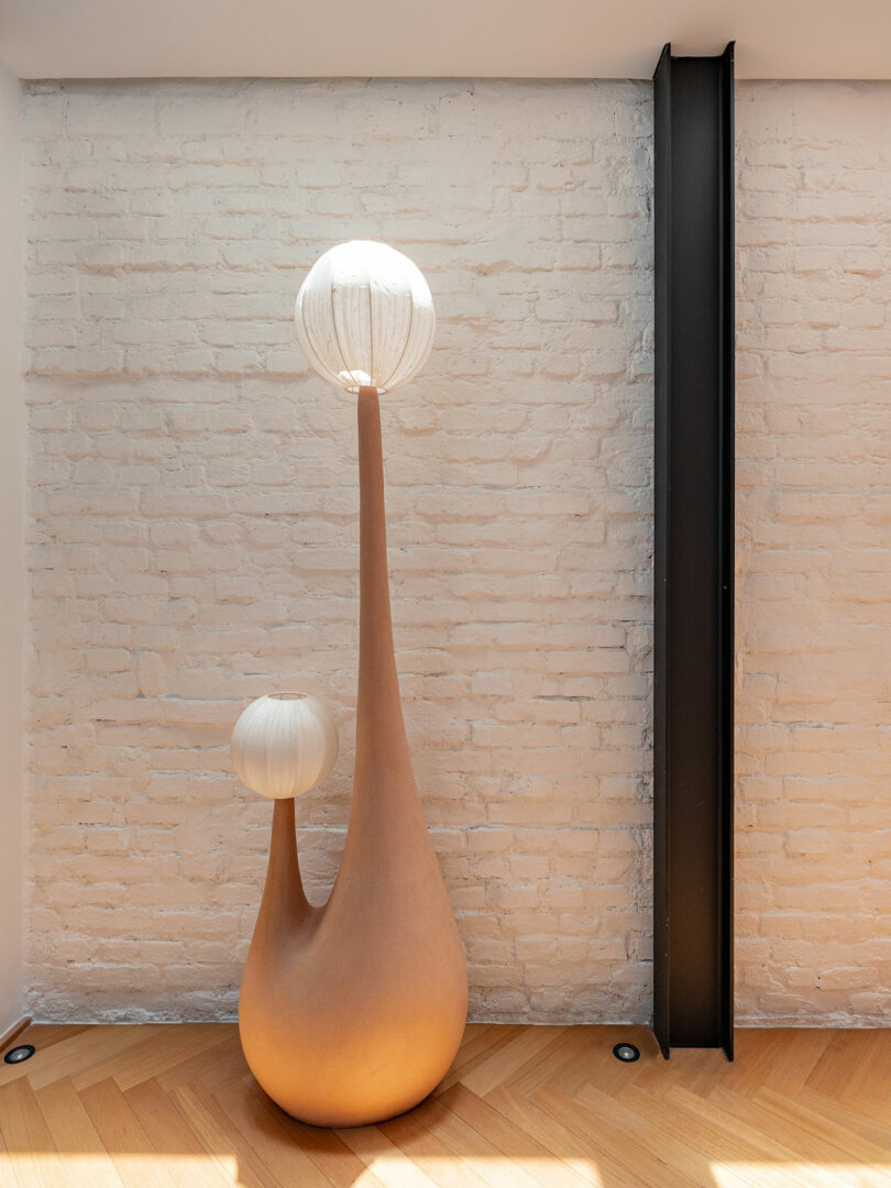 Tall sculptural vase with two elongated branches, each topped with a white textured sphere, placed against a white brick wall and wooden floor.