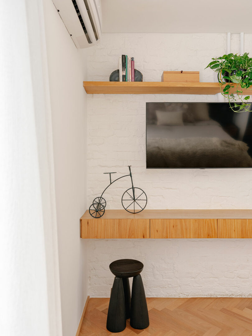 Minimalist corner with a wall-mounted TV, wooden shelves displaying books, a metal bicycle decor, a potted plant, and a small black stool on a light wood floor.