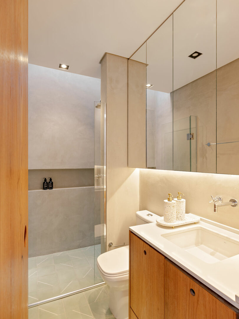 Modern bathroom with a glass shower enclosure, wall-mounted toilet, wooden vanity with sink, under-cabinet lighting, and large mirrored cabinets.