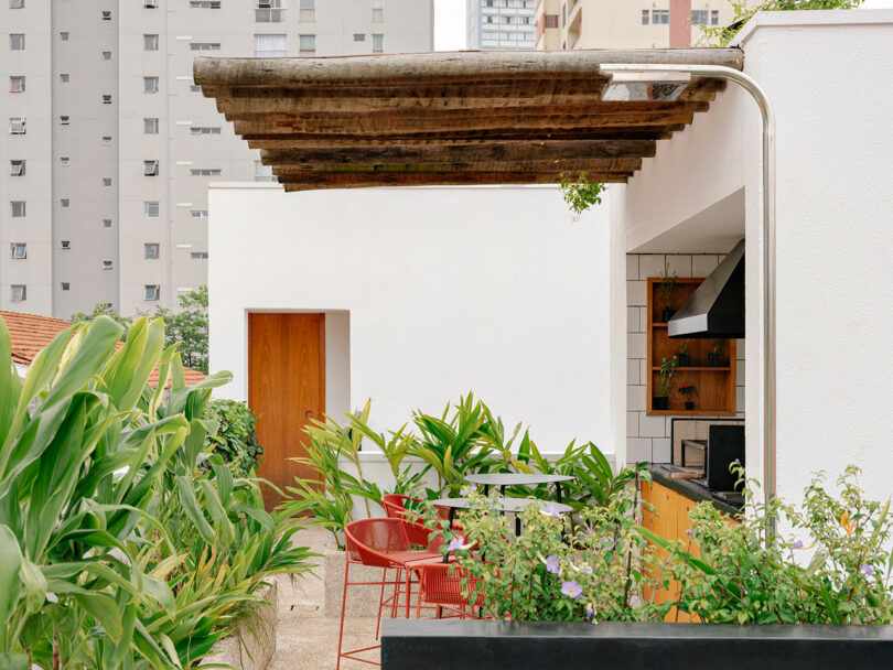 Rooftop patio with wooden pergola, red chairs, green plants, and a small outdoor kitchen against a white building; city apartment buildings in the background.